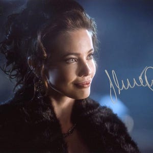 Puede incluir: Retrato de una mujer con cabello oscuro y abrigo de piel. La imagen presenta una firma en tinta plateada. La mujer mira hacia un lado con una ligera sonrisa. El fondo está borroso con tonos azules y blancos.