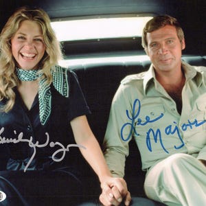 May include: A signed photograph of two people, a woman wearing a black shirt with a white and black scarf and a man wearing a beige shirt. The woman's signature is in silver ink and the man's signature is in blue ink. The signatures read "Jamie Lee Curtis" and "Lee Majors".