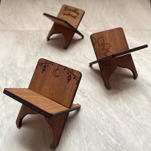 Op de afbeelding: Drie houten boekenstandaards met decoratieve ontwerpen. Eén standaard heeft de tekst "Ramadan Mubarak", een andere een bloemmotief en de derde een maan en lantaarns. De standaards zijn gemaakt van lichtbruin hout.