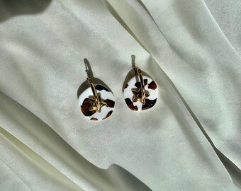 May include: A pair of hoop earrings featuring irregular, rounded white charms with brown cow-print spots. Each charm is accented with a small, gold-toned star-shaped metal charm attached to the hoop, creating a distinct contrast against the white and brown pattern.