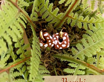 May include: A pair of square-shaped earrings featuring a brown base with white spots, suspended from gold-toned metal hoops. The earrings are positioned among green fern leaves, and a small rectangular label displays the text Bambi.