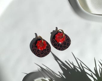 May include: A pair of hoop earrings featuring a circular brown pendant with a black leopard print pattern. Each earring is accented with a small, bright red, flower-shaped bead attached to a gold-toned metal hoop, creating a layered design.