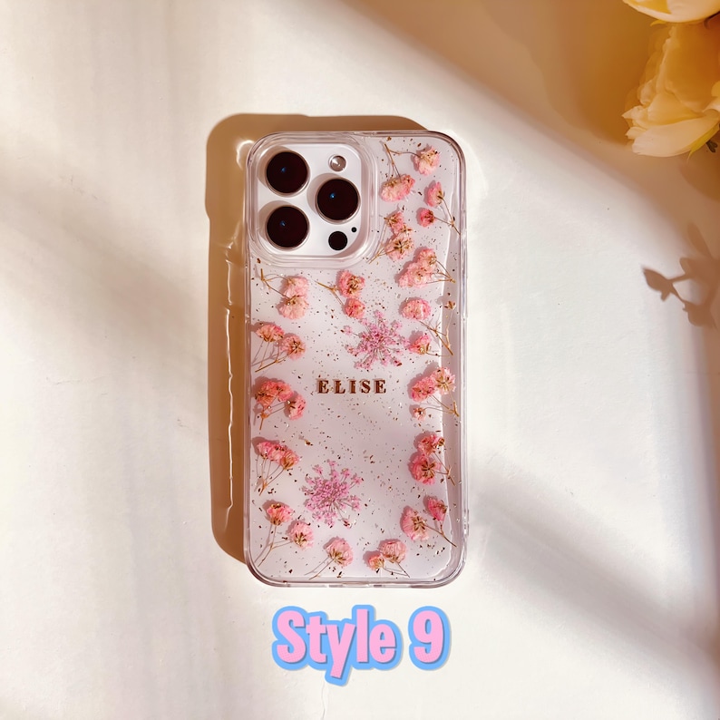 May include: A clear phone case with a floral design and the name "Elise" printed in gold. The case is filled with pink pressed flowers and glitter.