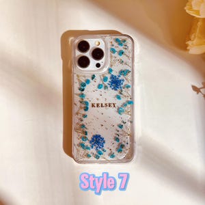 May include: Clear phone case with a floral design featuring blue flowers and gold glitter. The case has a personalized name "Kelsey" on it. The case is labeled "Style 7".
