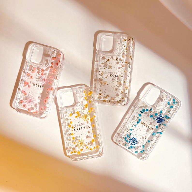 May include: Four clear phone cases with pressed flowers and gold lettering. The cases are pink, yellow, blue, and white with the names "Elise", "Kaylena", "Kedsen", and "Cecelia" respectively.