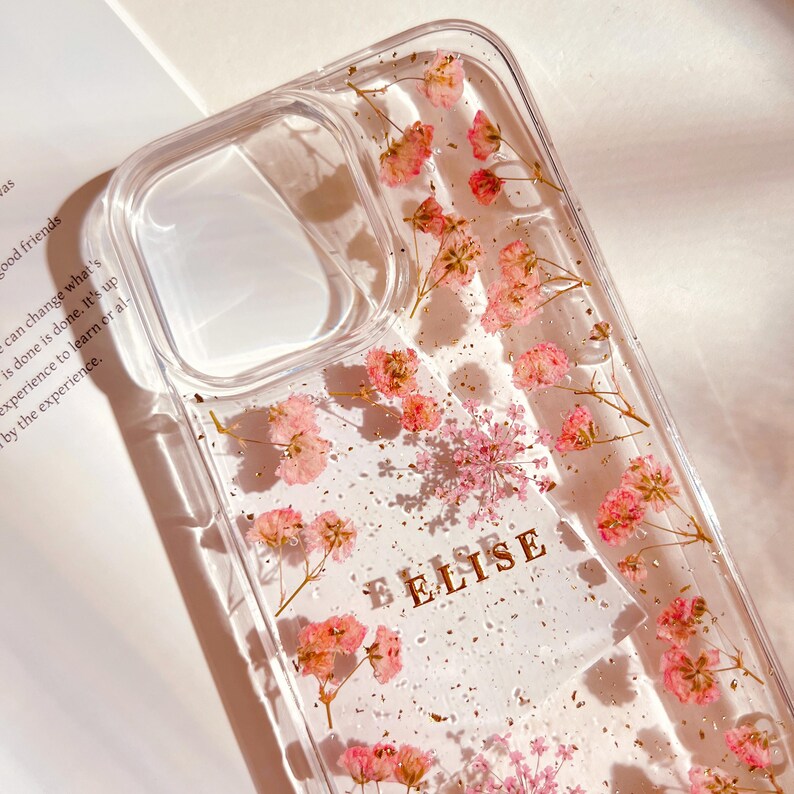 May include: A clear phone case with a floral design. The case has pink flowers and gold glitter embedded in the clear plastic. The word "FELISE" is written in gold letters on the case.