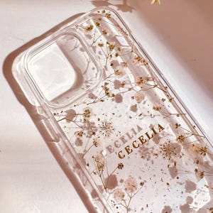 May include: A clear phone case with dried white flowers and gold glitter embedded inside. The name "CECELIA" is written in gold lettering on the case.