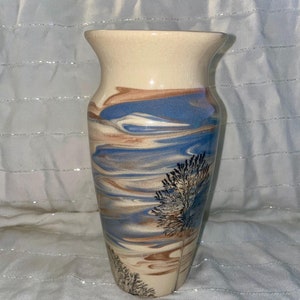 May include: A decorative ceramic vase with a cream-colored top and a flared rim. The body features a swirling design in shades of blue, brown, and cream, with a painted tree motif. The vase is approximately 8 inches tall.