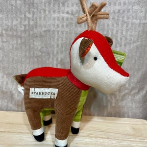 May include: A plush reindeer toy with a red head and back, brown body, and green legs. The reindeer has a white face, brown antlers, and a red ribbon around its neck. The Starbucks logo is on the side.