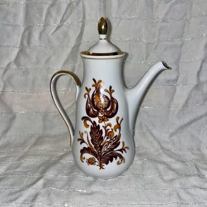 May include: White ceramic coffee pot with a lid and a curved spout. The pot features a brown and gold floral design and gold trim. The handle and lid finial are also gold. The pot is likely vintage and suitable for serving coffee or tea.