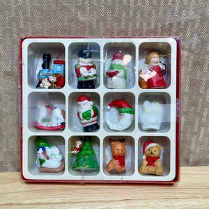 May include: A set of twelve Christmas ornaments in a red and white box. The ornaments include a train, snowman, angel, Santa Claus, and a teddy bear. The ornaments are painted in various colors.