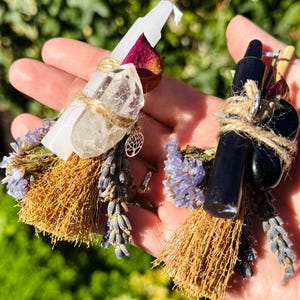 May include: Two miniature broomstick ornaments bound with twine, featuring dried lavender, rose petals, and crystals. One broom has a white candle and clear quartz, while the other has a black candle and a smooth black stone, both with a tree charm.