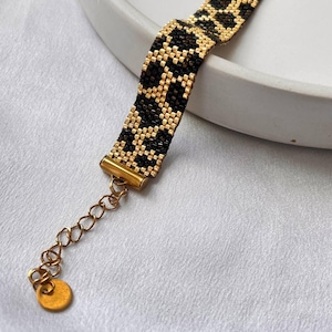 May include: A beaded bracelet with a leopard print design. The bracelet features black and gold beads, with gold-colored metal hardware and an adjustable chain. The bracelet is displayed on a white surface.