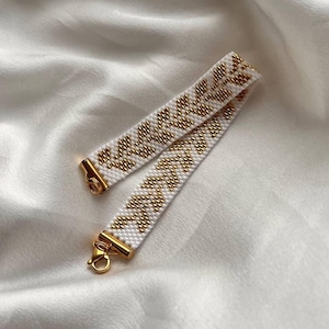 May include: White beaded bracelet with a gold leaf pattern and gold-colored clasps. The bracelet is made of small white beads with a repeating design of gold beads. The clasp is a simple hook and eye closure.