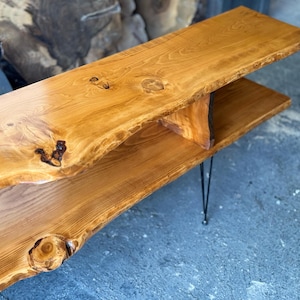 K&ouml;nnte beinhalten: Ein Holz-Couchtisch mit einer Live-Edge-Oberfl&auml;che und einem unteren Regal. Der Tisch hat eine nat&uuml;rliche Holzoptik und wird von schwarzen Haarnadelbeinen getragen.