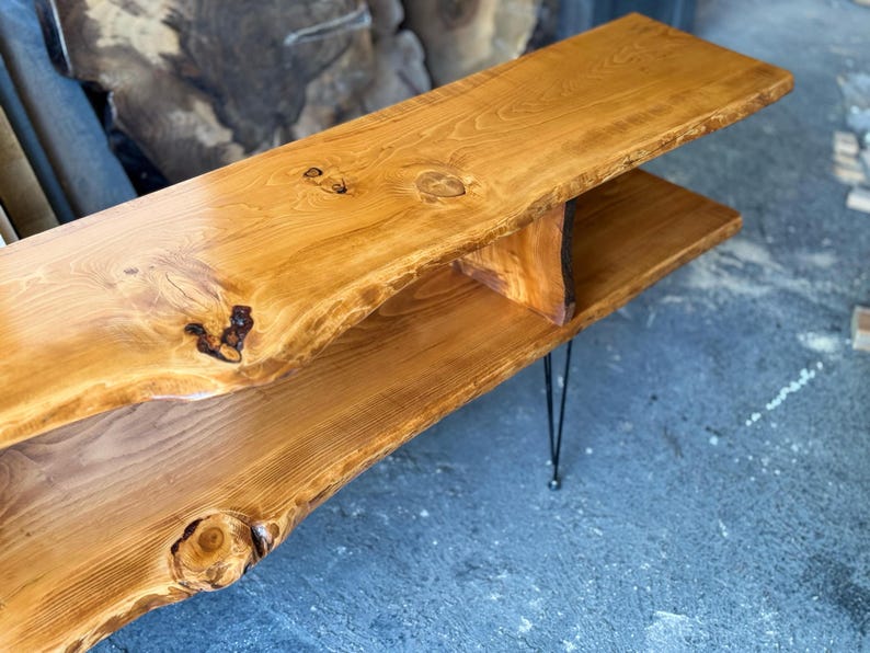K&ouml;nnte beinhalten: Ein Holz-Couchtisch mit einer Live-Edge-Oberfl&auml;che und einem unteren Regal. Der Tisch hat eine nat&uuml;rliche Holzoptik und wird von schwarzen Haarnadelbeinen getragen.