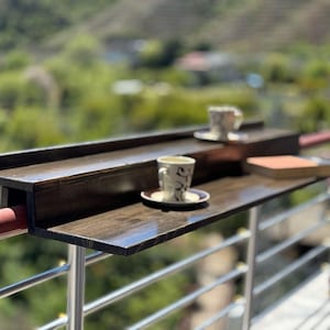 May include: A dark brown wooden table attached to a metal railing. The table has two shelves and is perfect for enjoying a cup of coffee or tea outdoors.
