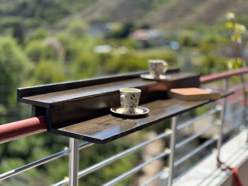 May include: A dark brown wooden table attached to a metal railing. The table has two shelves and is perfect for enjoying a cup of coffee or tea outdoors.