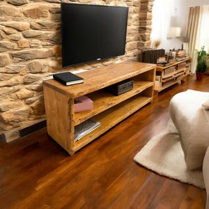 May include: A rustic, wooden TV stand with three shelves. A black TV is mounted on the stone wall above the stand. The stand holds a black book, a pink box, and a small black electronic device. The room has hardwood floors and a beige rug.