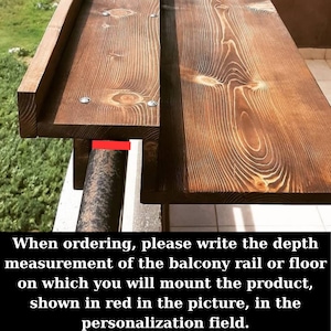 May include: A wooden shelf with a metal bracket, designed to be mounted on a balcony railing or floor. The shelf is made of natural wood with a rustic finish. The metal bracket is shown in red in the image.