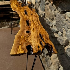 May include: A long, rustic wooden table with a live edge and natural wood grain. The table is supported by four black hairpin legs. The table is made of olive wood and has a natural, rustic finish.
