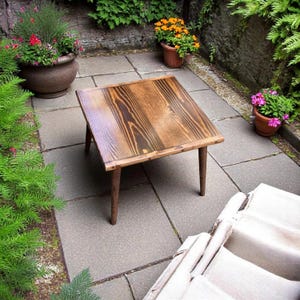 Peut inclure: Une table basse carrée en bois avec une finition en bois brun foncé et quatre pieds. La table est placée sur un patio avec des dalles grises.