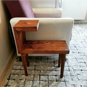 May include: A dark brown wooden side table with a unique L-shaped design. The table has a small shelf and two legs on each side. The table is sitting on a patterned rug.