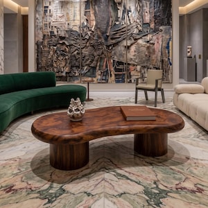 May include: A kidney-shaped coffee table made of dark brown wood with a smooth, rounded edge and two cylindrical pedestal bases. A small, textured, off-white ceramic sculpture and a rectangular brown book rest on the table's surface.
