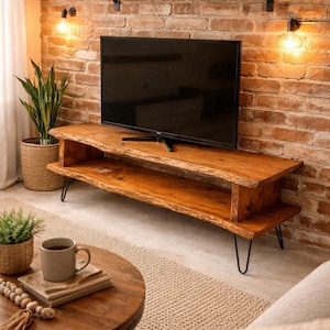 May include: A rustic wooden TV stand with a live edge design, supported by black hairpin legs. The stand is placed against a brick wall, with a black flat-screen TV on top. The room is decorated with plants and warm lighting.
