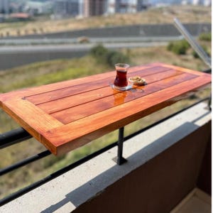 Op de afbeelding: Een rechthoekige houten balkontafel met een rijke, roodbruine afwerking. De tafel is gemonteerd op een zwarte metalen reling en bevat een glas thee en een klein schaaltje met noten. De tafel is ongeveer 76 cm lang.