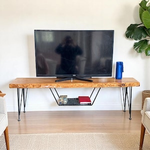 Peut inclure: Un meuble TV en bois avec un plateau en bois brut et des pieds en épingle à cheveux en métal noir. Le meuble dispose d'une étagère inférieure pour le rangement.