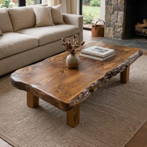 May include: A rectangular wooden coffee table with a natural edge and four short legs. The table is a warm brown color and sits on a beige rug. A small vase with dried flowers and a stack of books are on the table.