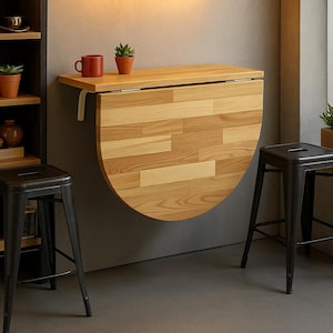 May include: A wall-mounted, folding wooden table with a light wood grain pattern. The table is half-circle when folded down. Two black metal stools are in front of the table. A red mug and a small potted plant sit on the table.