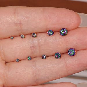 May include: An assortment of iridescent stud earrings in different sizes. The earrings exhibit a spectrum of colours, including green, blue, and purple, set against a neutral backdrop. The earrings are round and seem to be made of a reflective material.