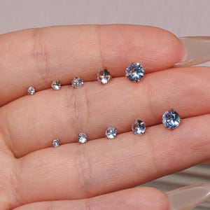 May include: A collection of small, round stud earrings in varying sizes. The earrings are clear and light blue, with a sparkling, reflective quality. The earrings are displayed on a hand.