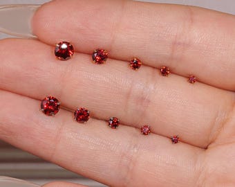 Può includere: Una collezione di orecchini a lobo con gemme rosse di varie dimensioni. Gli orecchini sono rotondi e sfaccettati, che mostrano un colore rosso vibrante. Gli orecchini sono disposti in due file, mostrando le variazioni di dimensione.