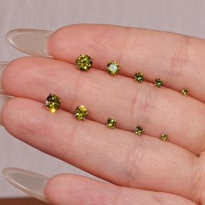 May include: A collection of small, round, olive-green gemstone stud earrings of varying sizes. The earrings are displayed on a light-toned background, showcasing their color and sparkle. The gemstones are set in a simple, classic style.