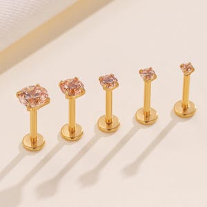 Op de afbeelding: Een verzameling van zes goudkleurige lip- of kraakbeen studs met licht perzik-kleurige cubic zirconia stenen. De studs zijn in een rij gerangschikt, waarbij de stenen in grootte afnemen. De achtergrond is lichtbeige.
