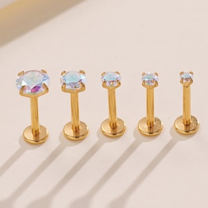 May include: Five gold-colored lip piercing studs with iridescent, round gemstones. The studs are arranged in a row, decreasing in size from left to right. Each stud has a flat base and a post.
