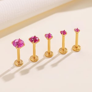 Puede incluir: Cinco piercings labiales dorados con gemas rosas de diferentes tamaños. Los piercings tienen una base plana y un poste que se extiende hasta la gema. Las gemas son de color rosa vibrante y están talladas en diferentes formas.