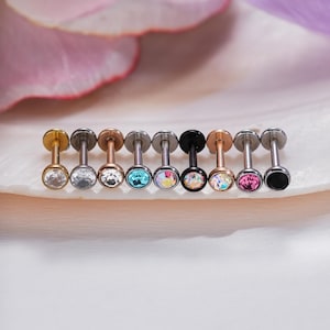 May include: A row of nine labret studs with flat back discs and circular front settings. The metal finishes include gold, silver, rose gold, and black. The front settings feature various round gemstones in clear, blue, iridescent, pink, and black.