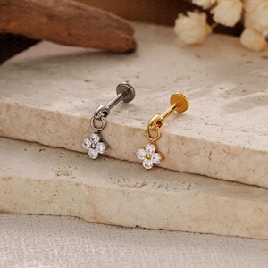May include: Two earrings, one silver-toned and one gold-toned, each featuring a small, dangling four-petal flower design. The flowers are adorned with clear, sparkling gemstones. The earrings are displayed on a textured, light-colored surface.