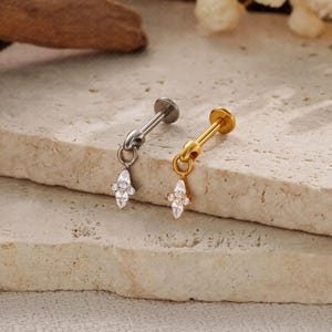 May include: Two ear piercing studs, one silver and one gold, each with a dangling, marquise-shaped charm. The charms are adorned with clear, sparkling crystals. The studs are displayed on a textured, light-coloured surface.