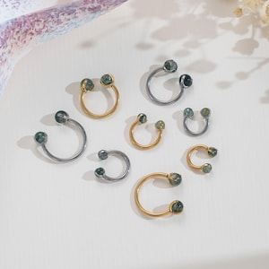 Può includere: Una collezione di sette bilancieri circolari a forma di ferro di cavallo in tonalità oro e metallo scuro. Ogni pezzo presenta due piccole perle di pietra rotonde color verde muschio alle estremità delle barre curve per i piercing.