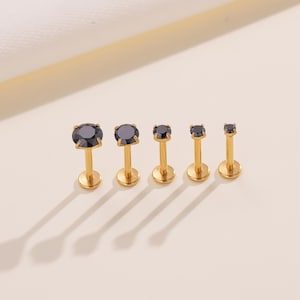 May include: Five gold-colored lip or cartilage studs with black gemstones. The studs are arranged in a row, each with a different size of black stone. The posts are straight and have a flat, round base.