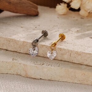 May include: Two pieces of jewelry, one silver and one gold, each featuring a small, clear, heart-shaped charm. The charms dangle from a post with a flat top, designed for ear piercings. The jewelry is set against a neutral, textured background.