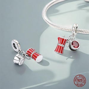 May include: Silver charm pendants featuring a red and white coffee maker and a coffee cup. One charm has a red gem. The charms are attached to a silver bracelet. The charms are made of 925 sterling silver.