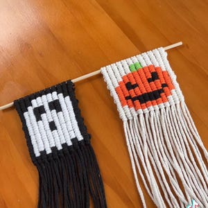 Könnte beinhalten: Halloween-Dekorationen aus Makramee mit einem pixeligen Geist und einem Kürbis-Design. Der Geist ist schwarz-weiß, der Kürbis orange, schwarz und grün. Jedes Stück hängt an einem Holzstab mit Fransen.