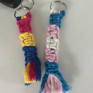 Op de afbeelding: Twee handgemaakte macramé sleutelhangers. De ene sleutelhanger heeft roze, gele, blauwe en roze secties. De andere sleutelhanger heeft blauwe, roze, witte en blauwe secties. Beide hebben zilveren sleutelringen en kleurrijke franjes.