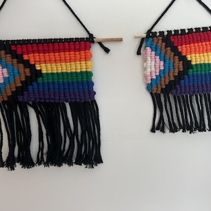 Op de afbeelding: Twee macramé wandkleden met regenboog- en zwarte franjes. Het grotere stuk heeft een regenboogverloop met een zwart chevronpatroon en een Progress Pride vlagontwerp. Het kleinere stuk heeft een vergelijkbaar ontwerp. Beide zijn opgehangen aan houten stokken.